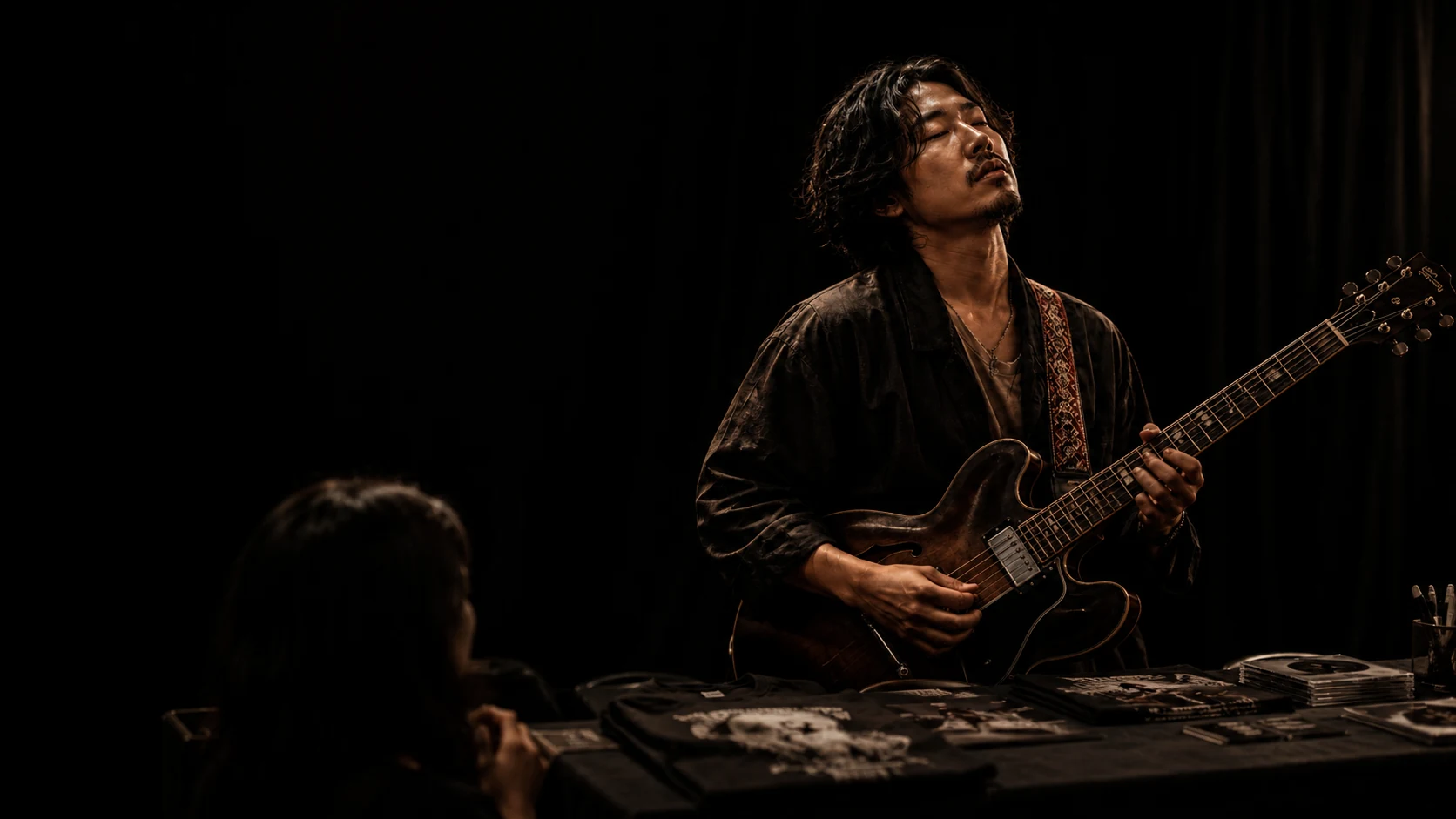 A guitarist plays with intensity behind a table of records and booklets.